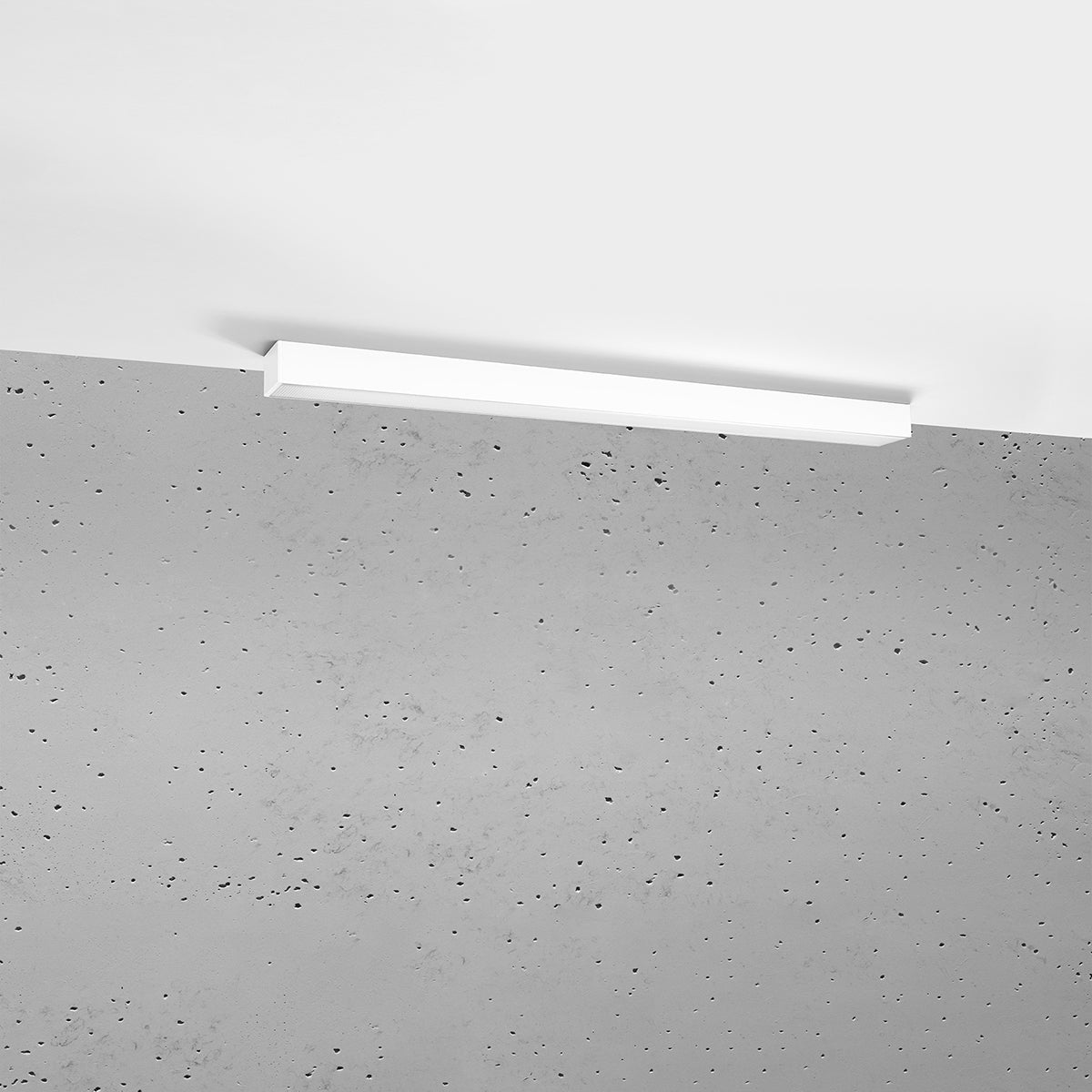Luminaire linéaire blanc fixé au plafond devant un mur béton brut, éteint