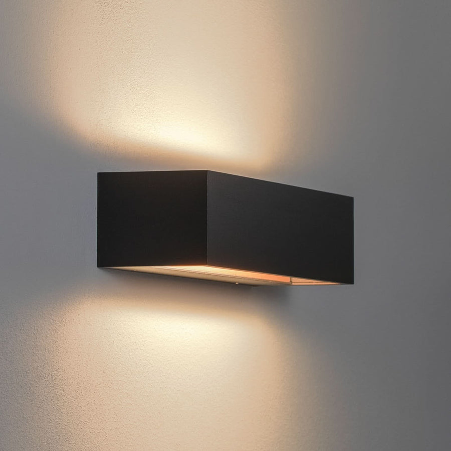 Applique murale noire allumée sur mur clair, lumière chaude diffusée vers le haut et le bas.