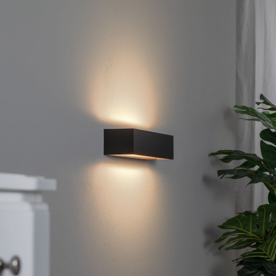 Applique noire de face sur mur gris, halo haut et bas, plante floue à droite.