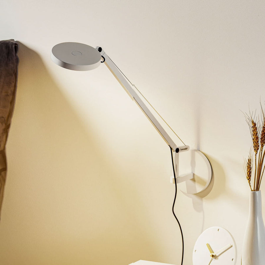 Applique murale blanche articulée éclairant un mur beige, près d’un tissu et d’épis.
