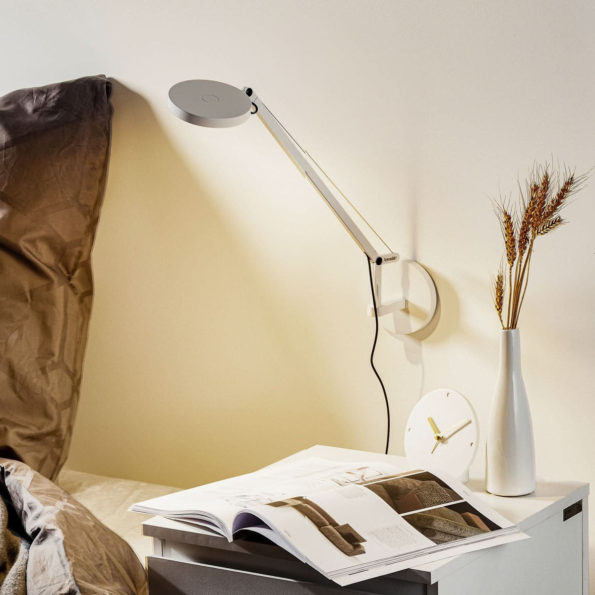 Vue de la lampe au-dessus d’un chevet, avec livre ouvert, horloge et vase.