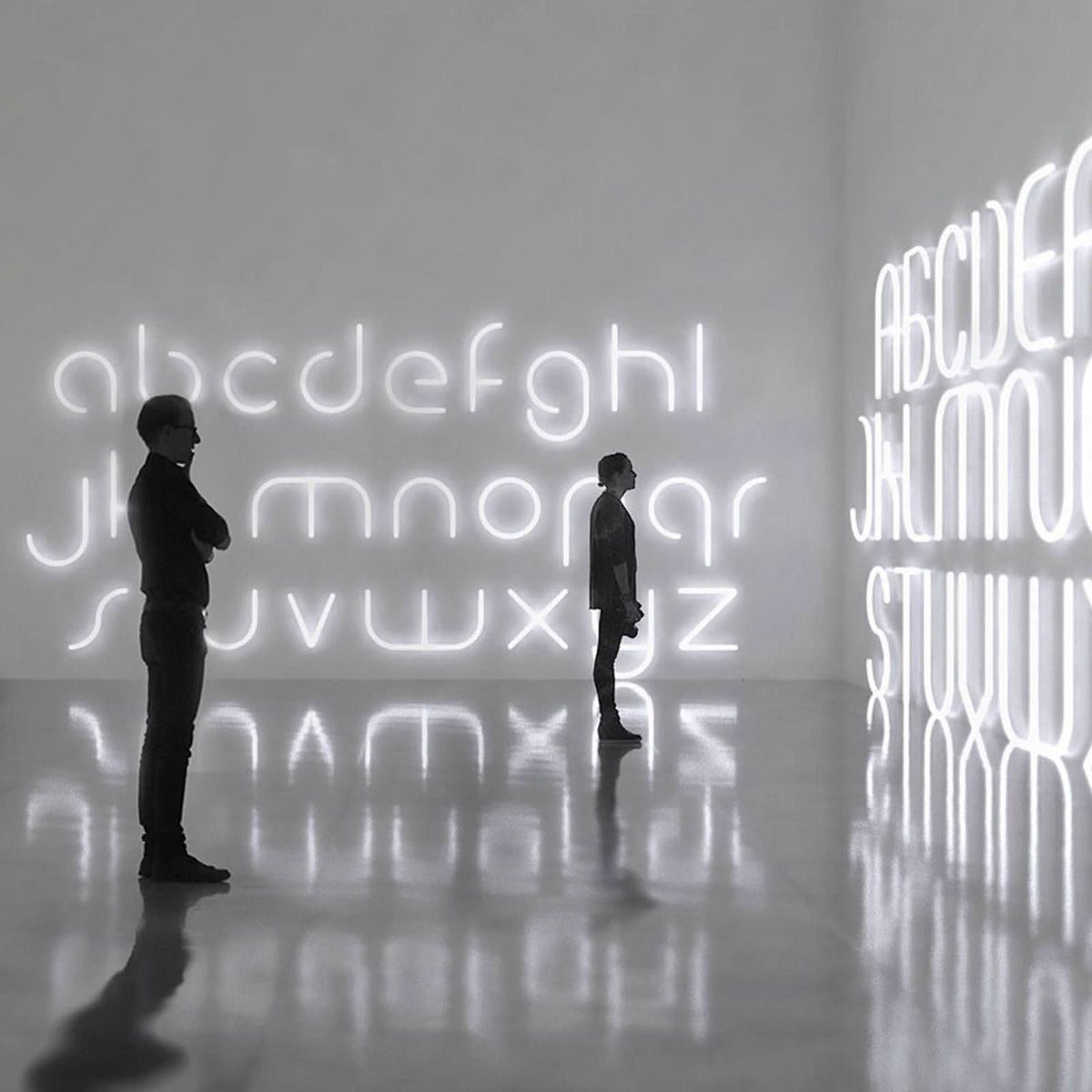 Deux personnes contemplent des lettres lumineuses blanches disposées sur un mur, reflet au sol.