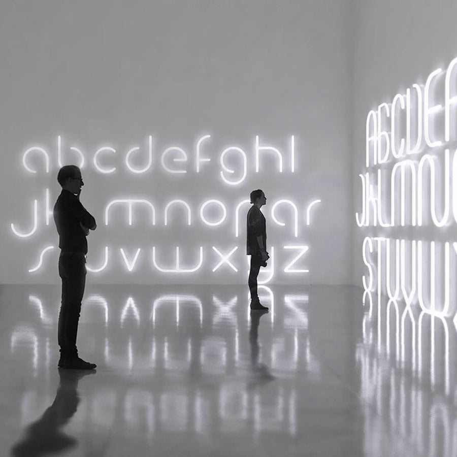 Deux personnes contemplent des lettres lumineuses blanches disposées sur un mur, reflet au sol.