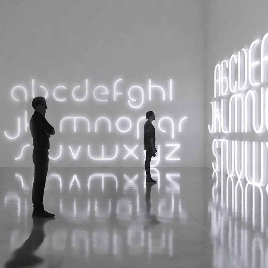 Deux silhouettes observent un mur couvert de lettres lumineuses blanches reflétées sur le sol.