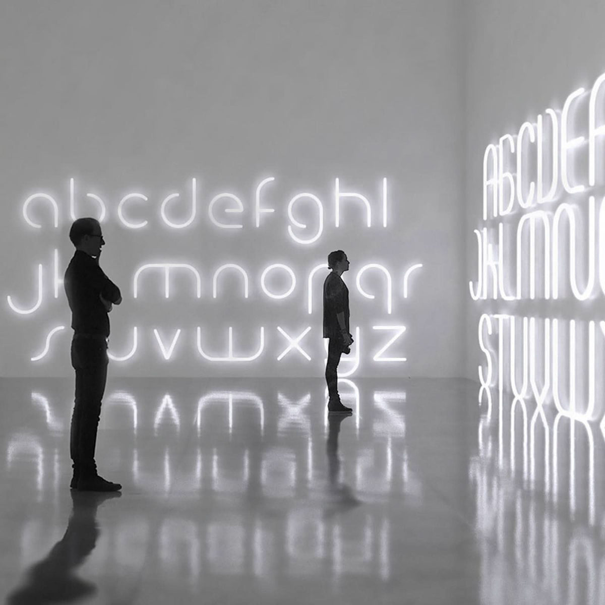 Deux silhouettes contemplent un alphabet lumineux blanc installé sur les murs d'une galerie.