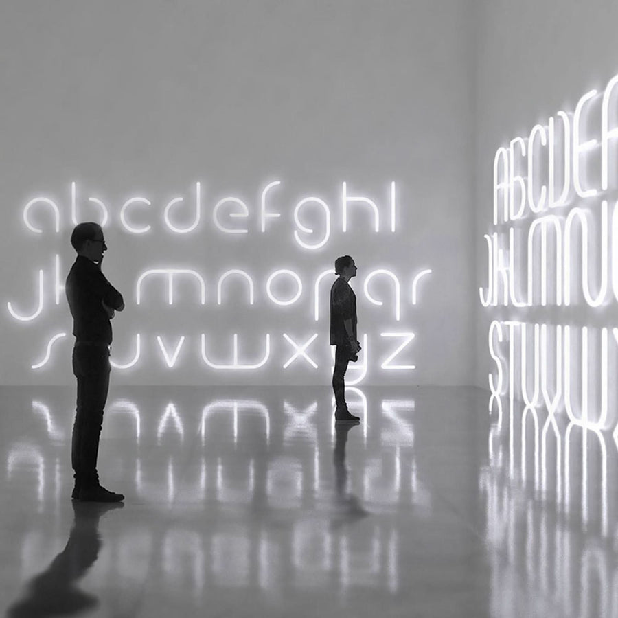 Deux silhouettes contemplent un alphabet lumineux blanc installé sur les murs d'une galerie.