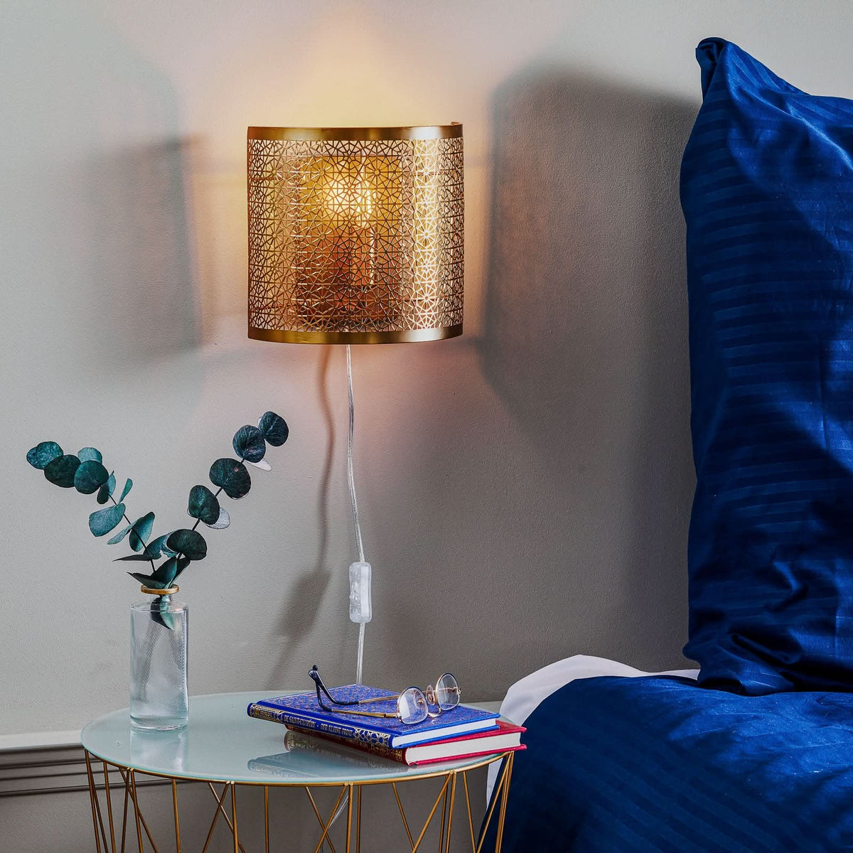 Applique en laiton allumée au-dessus d’une table de chevet, à côté d’un lit bleu.