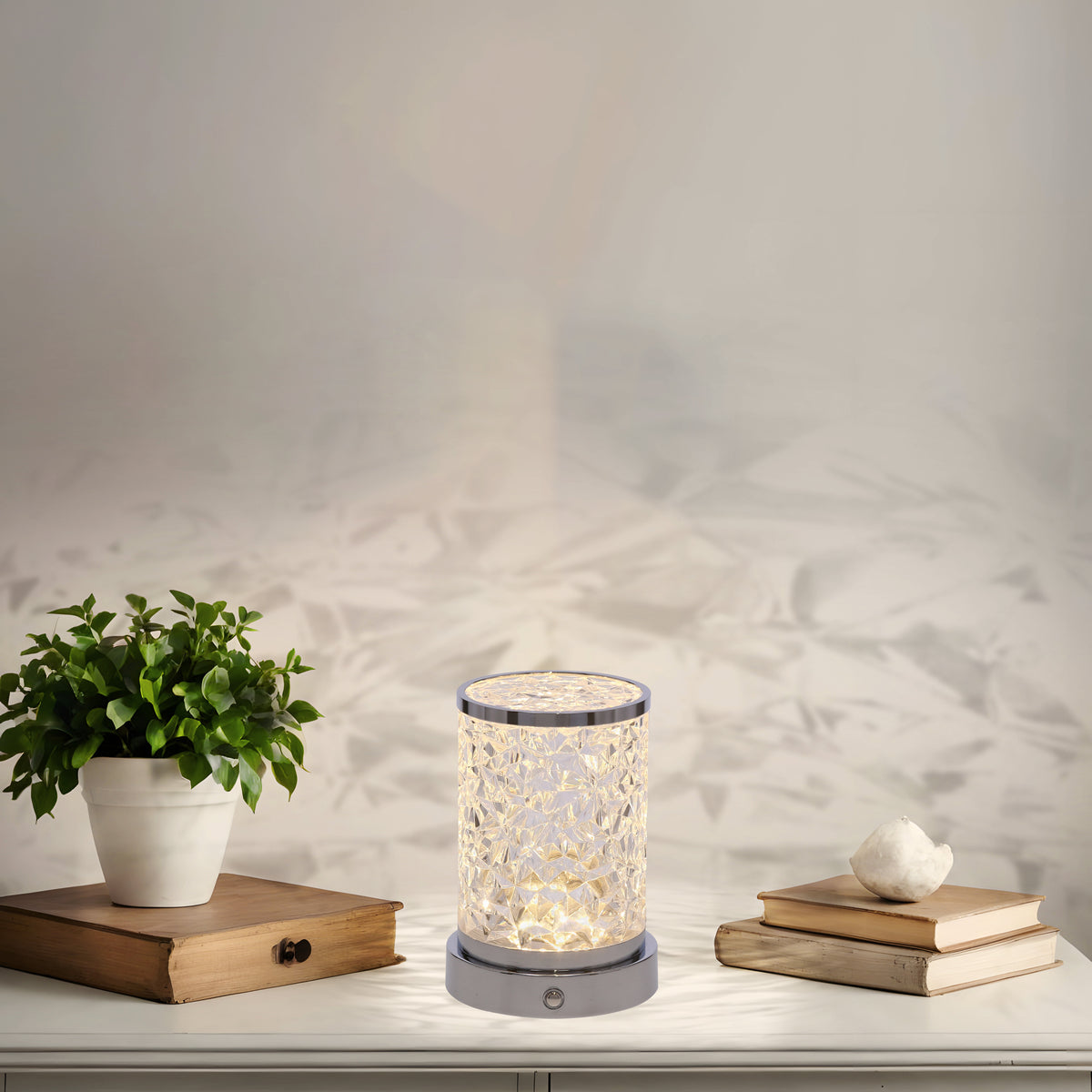 Lampe allumée posée sur meuble blanc, avec plante en pot et livres, ambiance chaleureuse.