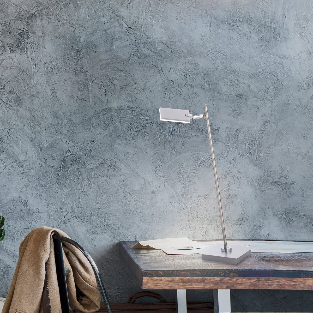 Mise en ambiance sur un bureau en bois, devant un mur gris texturé, manteau posé sur une chaise.