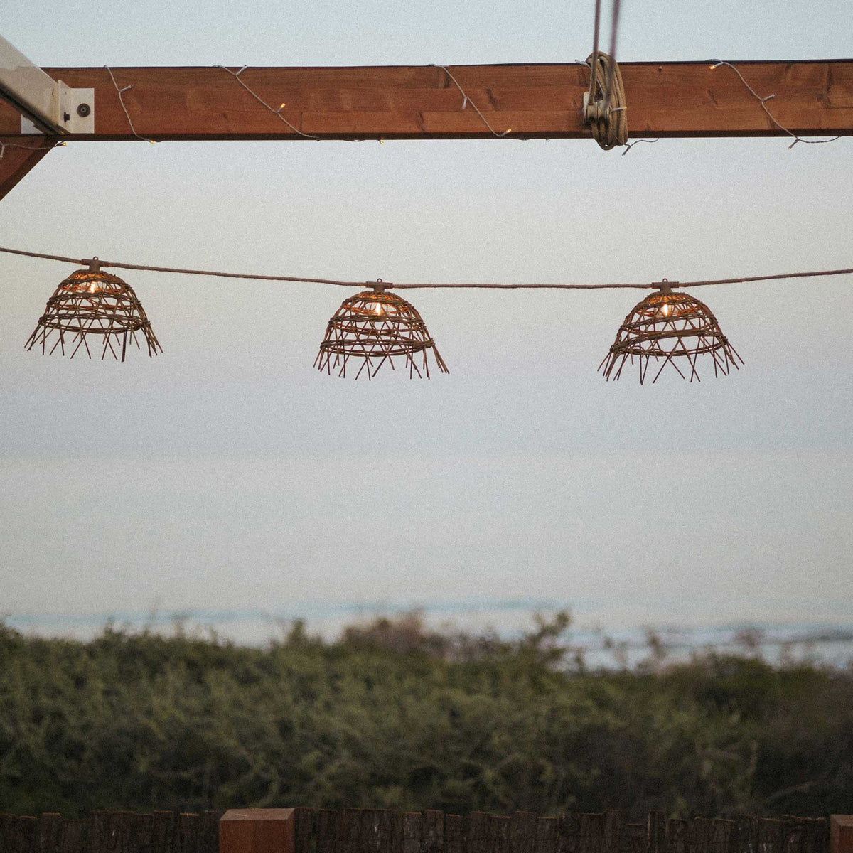 Trois abat-jours en rotin suspendus devant un paysage de mer et de collines lointaines.