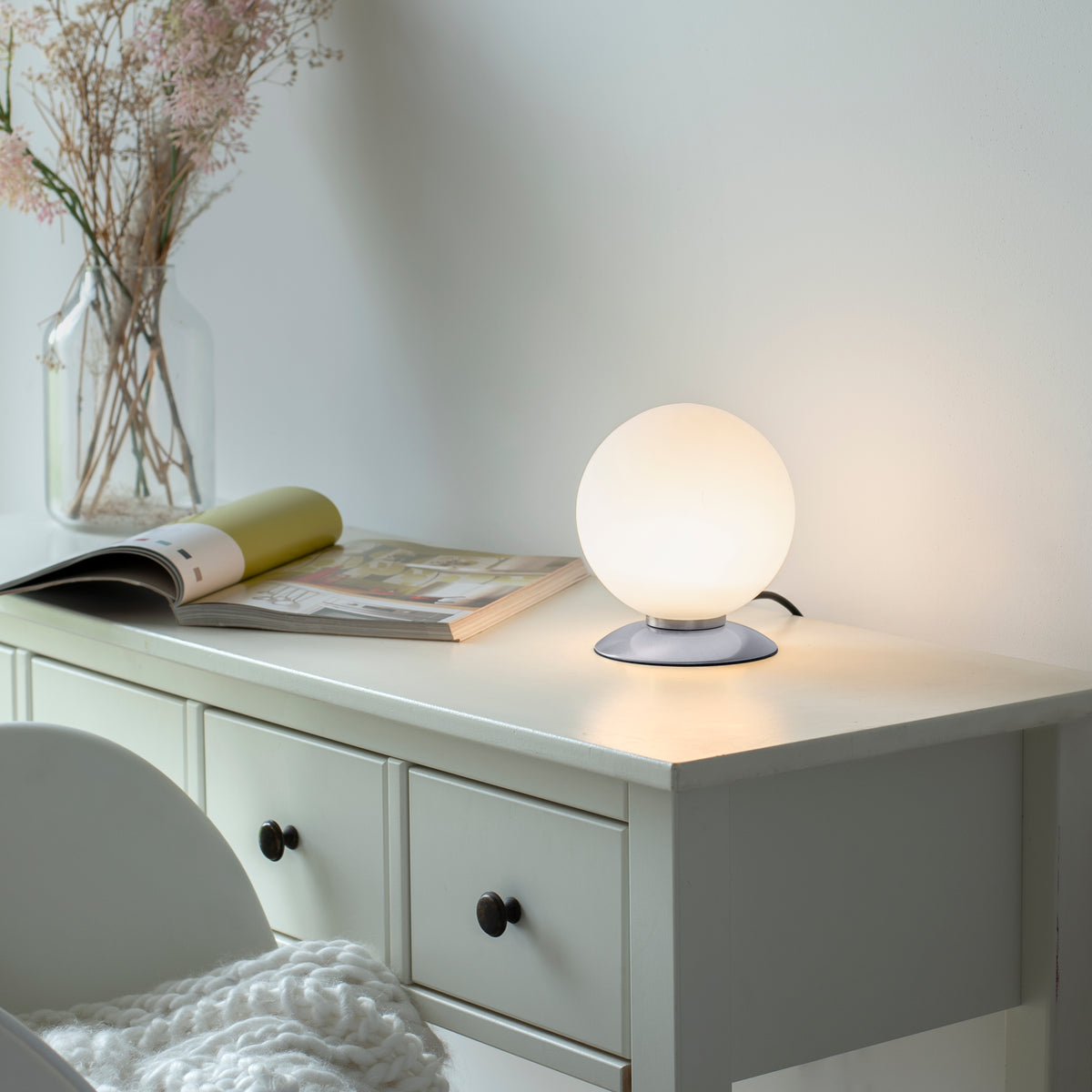 Lampe allumée posée sur une commode blanche, ambiance chaleureuse avec livre et vase.