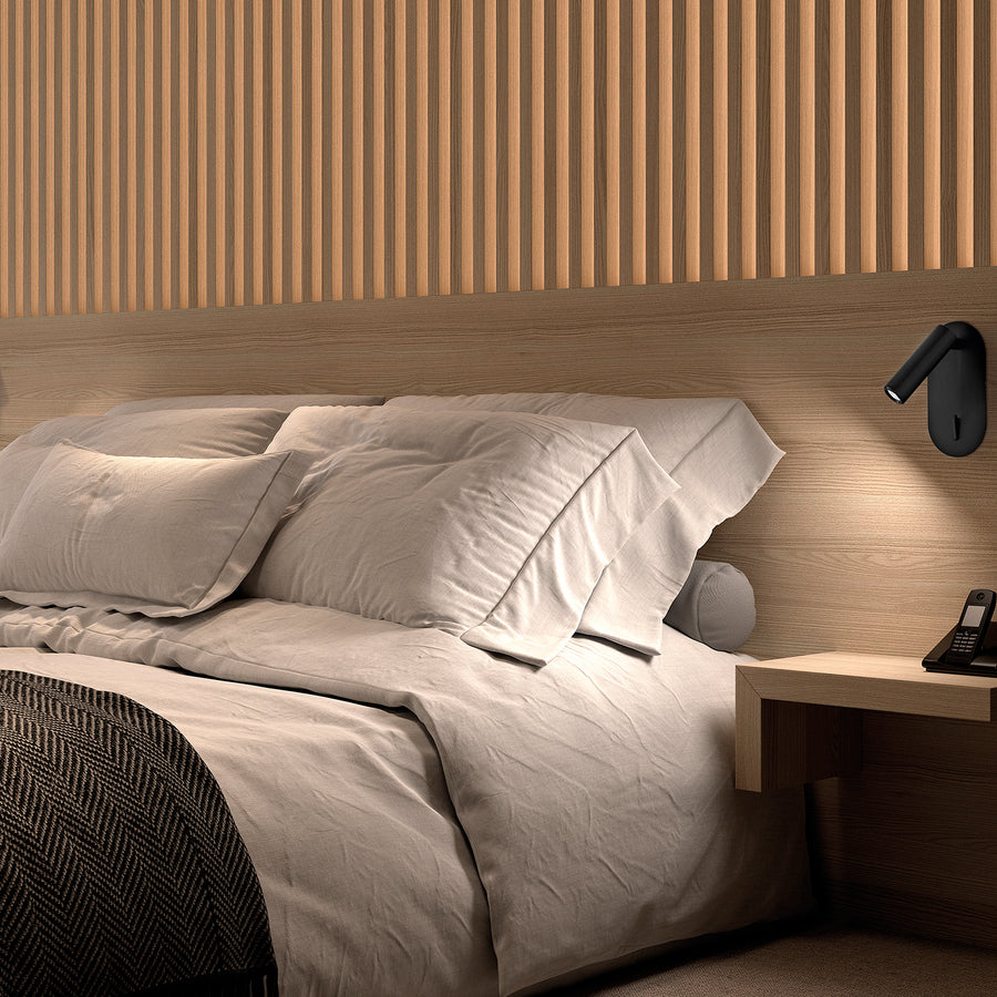 Mise en ambiance chambre en bois, applique noire éclairant la tête de lit et les coussins.