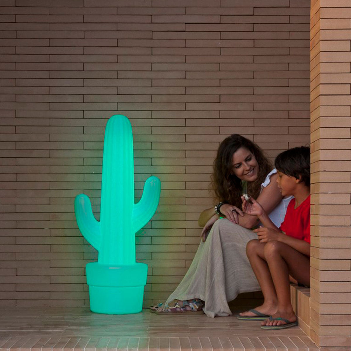 Femme et enfant près du lampadaire cactus vert lumineux, ambiance chaleureuse sur terrasse.
