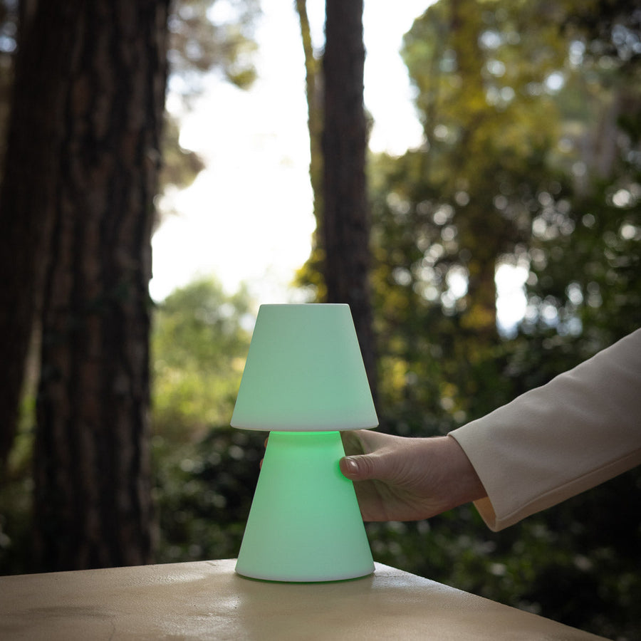 Une main actionne une lampe à poser verte allumée posée sur une table, devant des arbres.