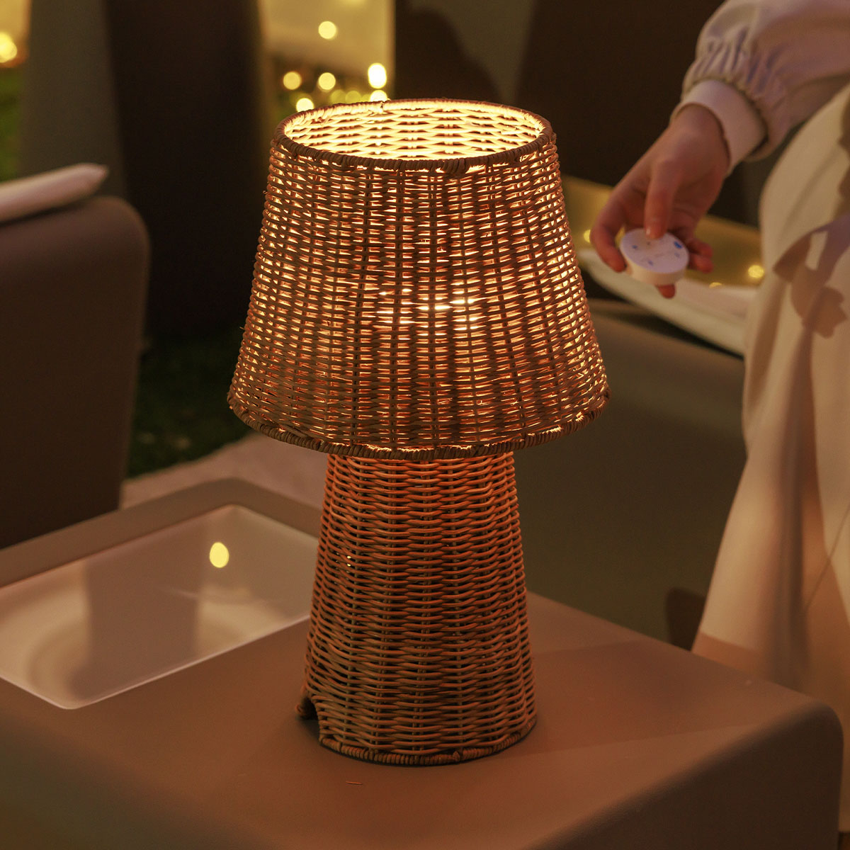 Lampe à poser en rotin tressé allumée sur table basse, main tenant une télécommande