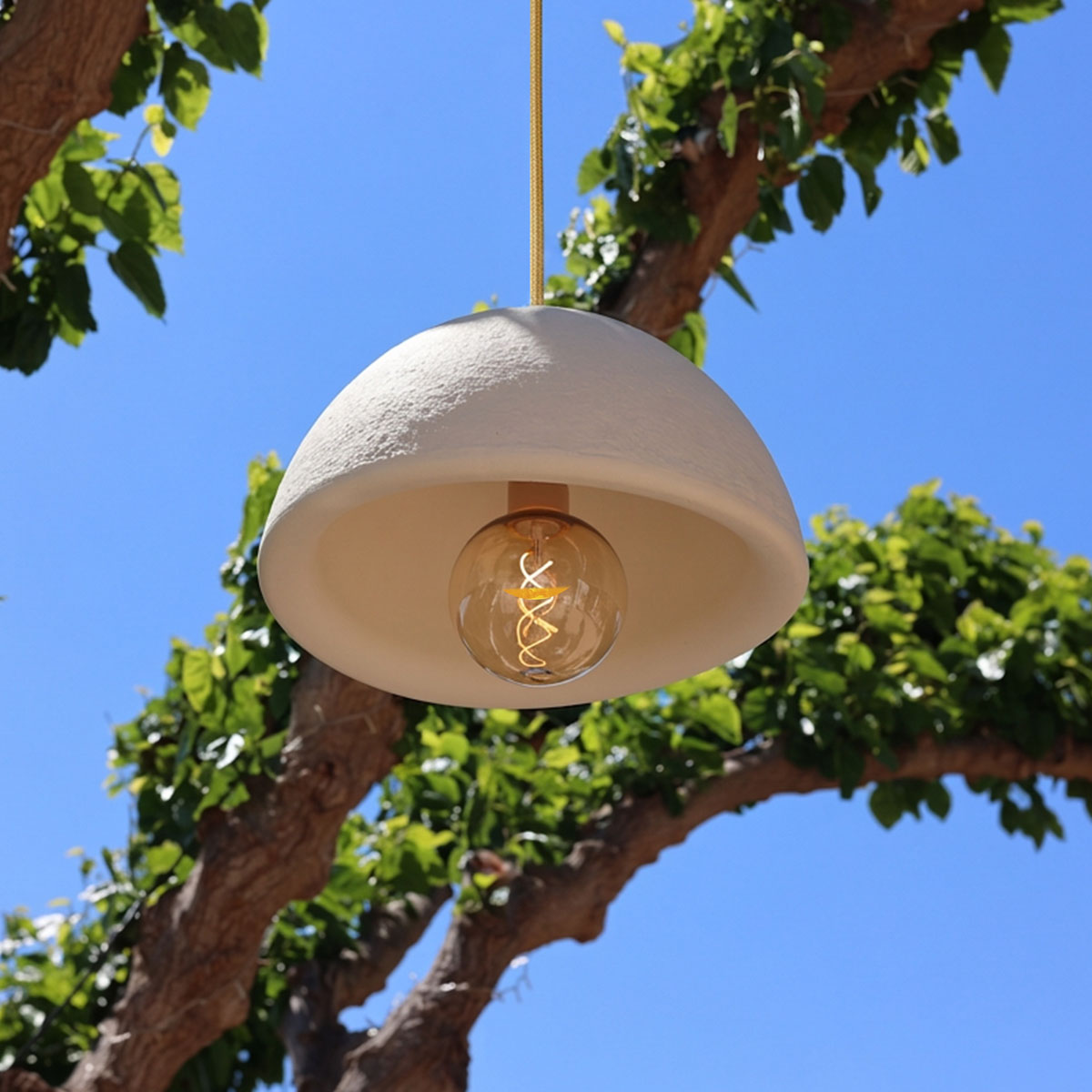 Suspension blanche sous un ciel bleu, accrochée à une branche avec feuillage vert, ampoule apparente.