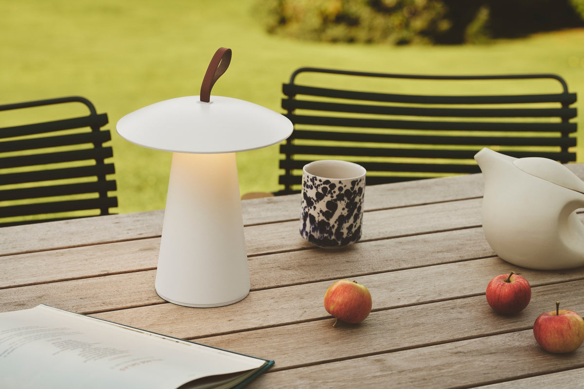 Lampe blanche sur table de jardin entourée de pommes, livre et théière.