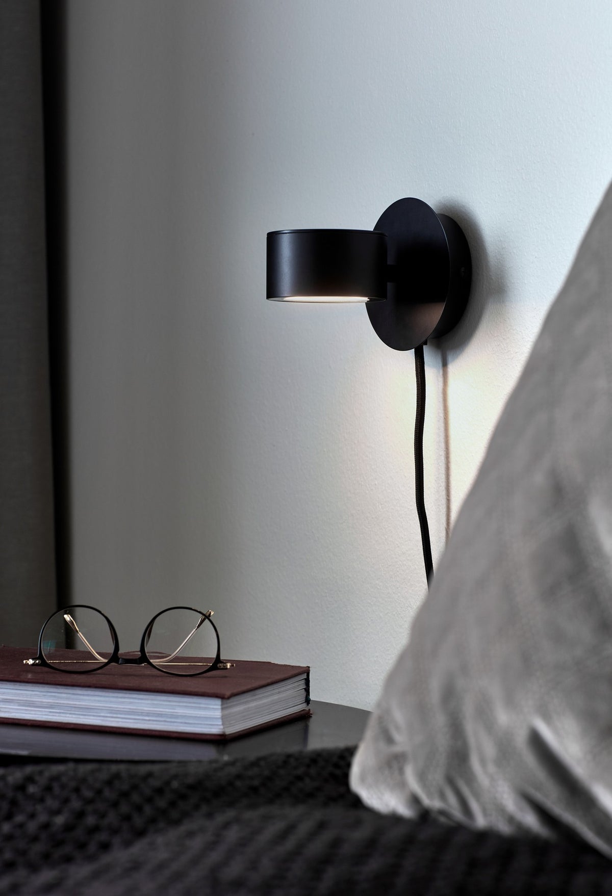 Mise en ambiance dans une chambre, l'applique éclaire un livre et des lunettes.