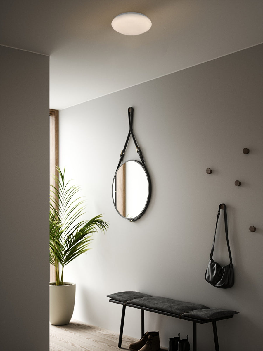 Ambiance entrée sombre, luminaire rond allumé éclairant le mur gris et le banc