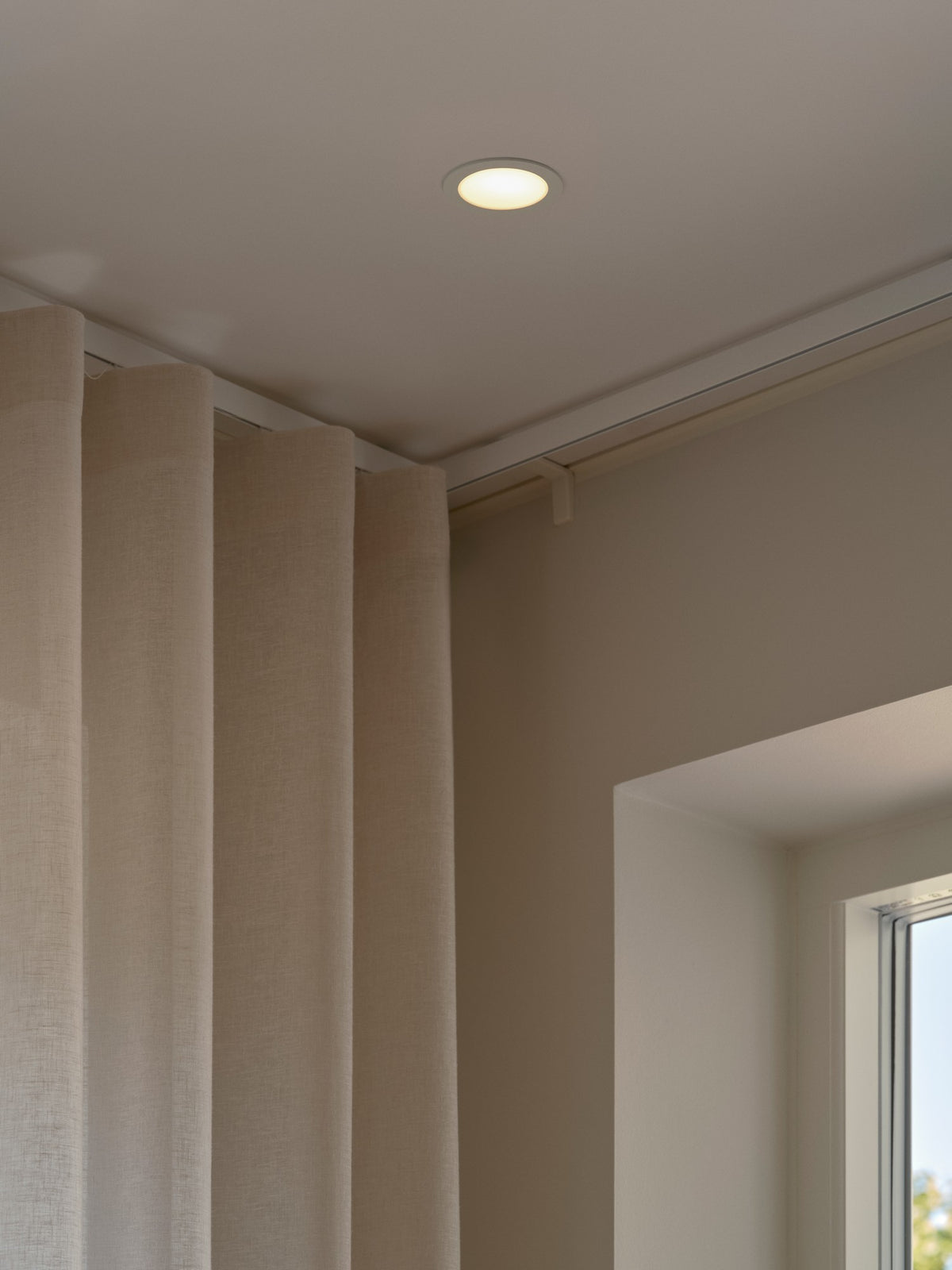 Mise en ambiance : spot encastré allumé au plafond près de rideaux beiges et fenêtre.