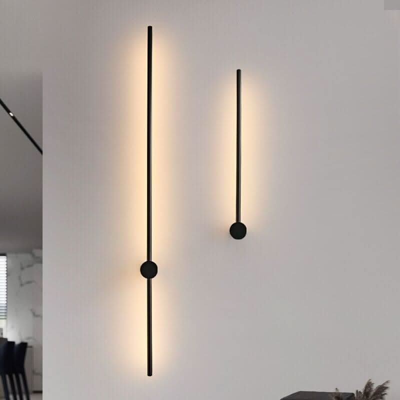 Deux appliques noires allumées, longueurs différentes, halos lumineux verticaux sur mur clair