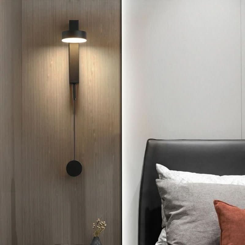 Applique murale noire allumée sur une paroi en bois, au-dessus d'un lit gris.