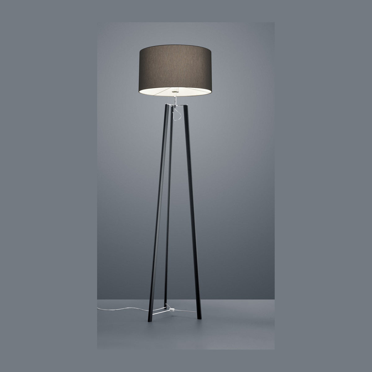 Lampadaire noir à trois pieds vu de face, abat-jour cylindrique gris taupe sur fond gris