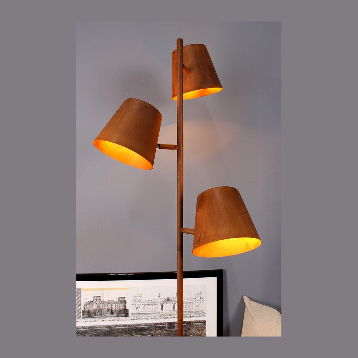 Lampadaire entier à trois abat-jours coniques rouille, posé sur parquet clair devant un mur gris.