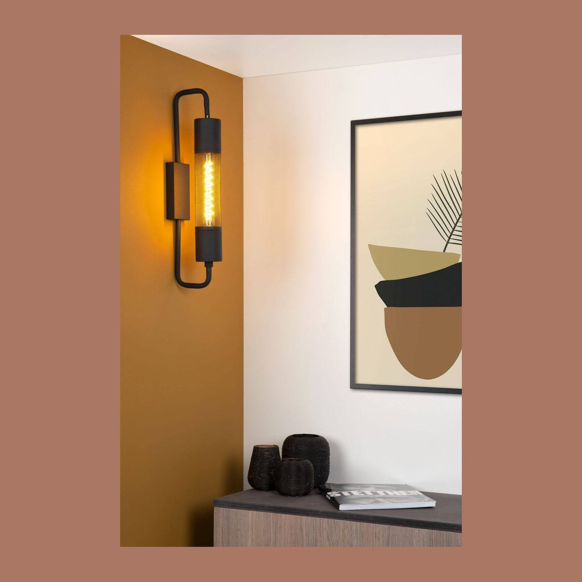 Mise en ambiance : applique murale noire allumée sur un mur ocre, près d'un cadre.