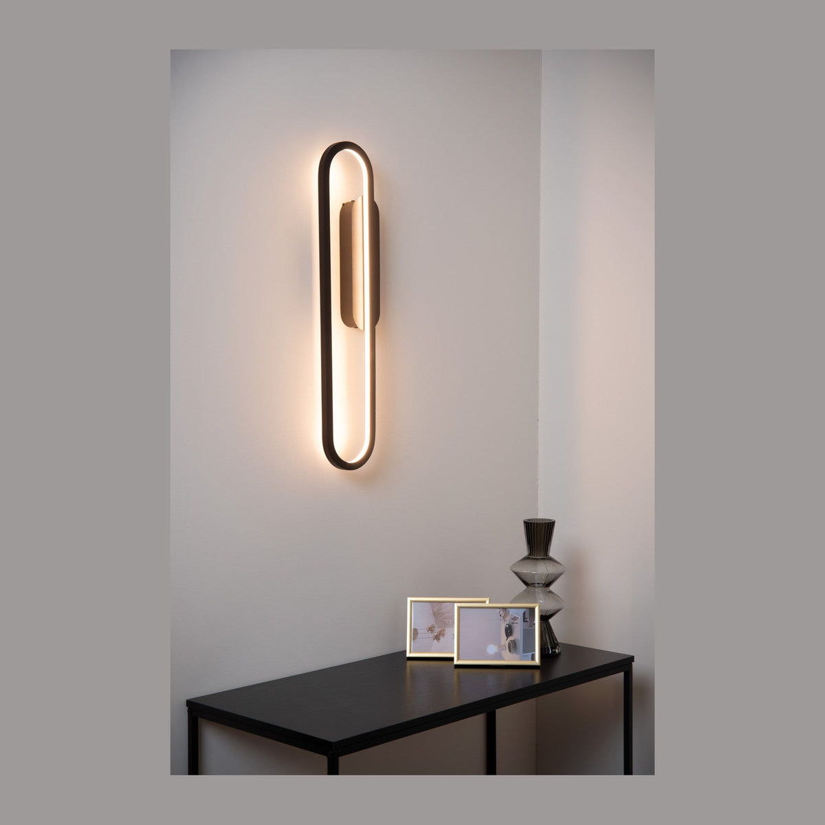 Mise en ambiance : applique ovale noire allumée sur mur clair, table noire et cadres photo
