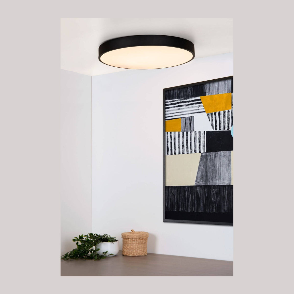 Plafonnier rond noir allumé au plafond dans un intérieur avec tableau et plante.