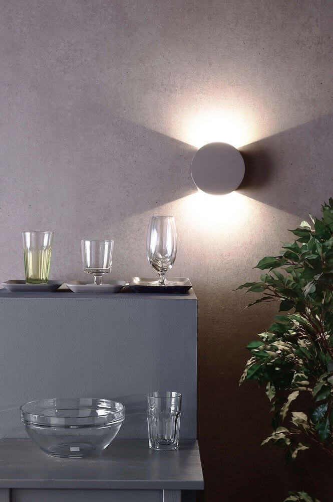 Applique murale allumée sur un mur gris, éclairage en croix et verres posés.