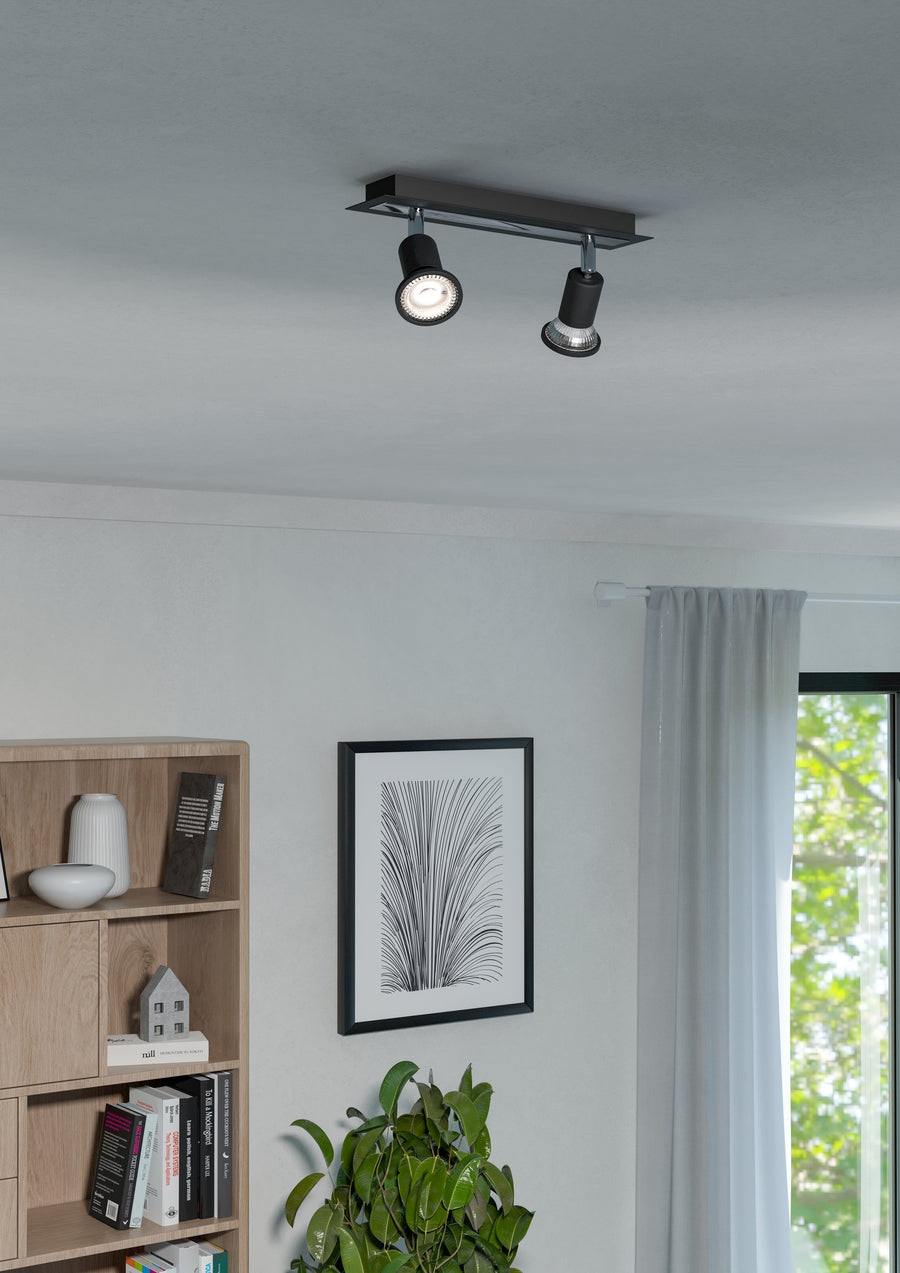 Spot noir allumé fixé au plafond dans un salon avec bibliothèque et plante.