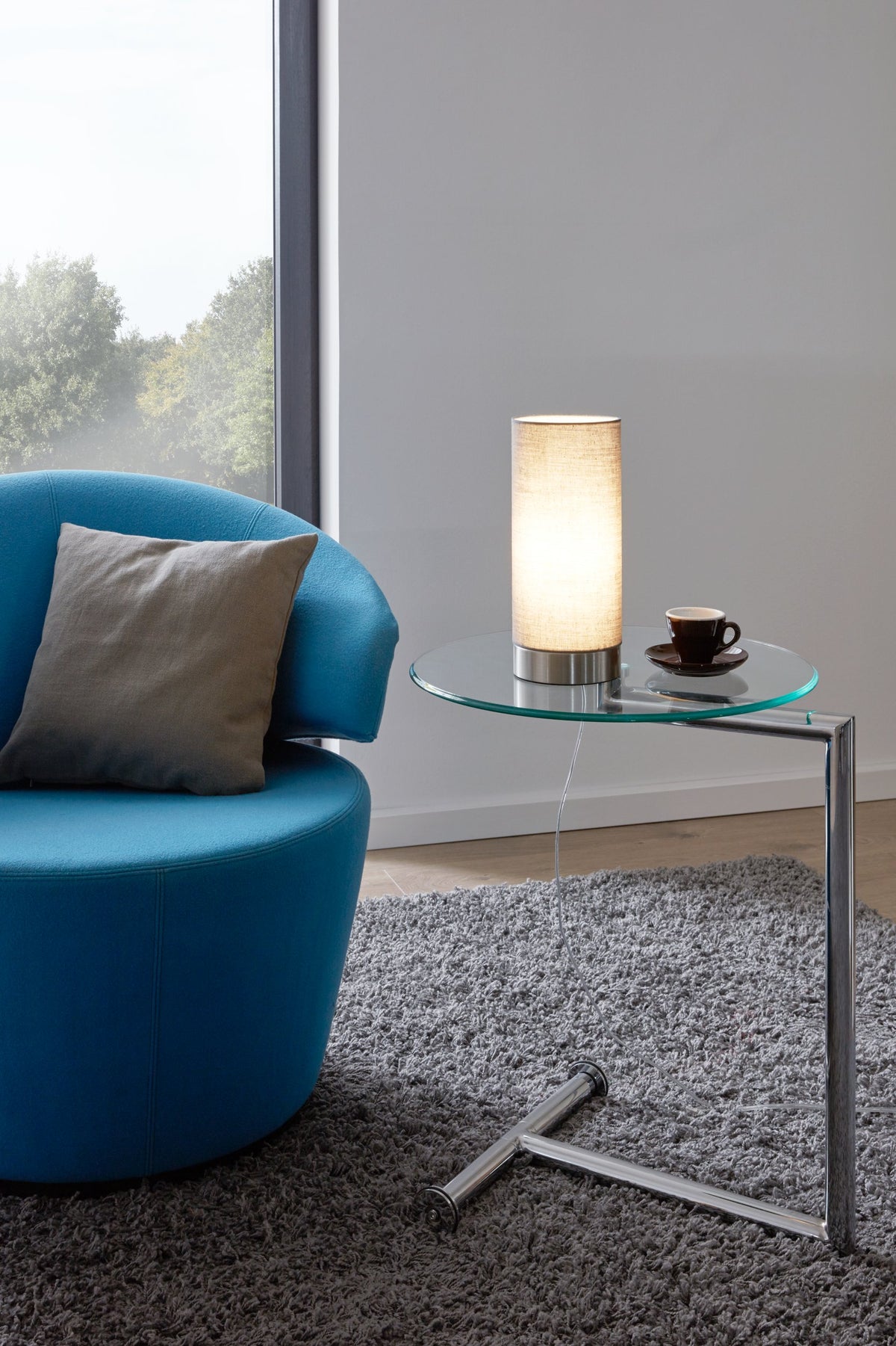 Mise en ambiance : lampe allumée posée sur table basse en verre près d'un fauteuil bleu