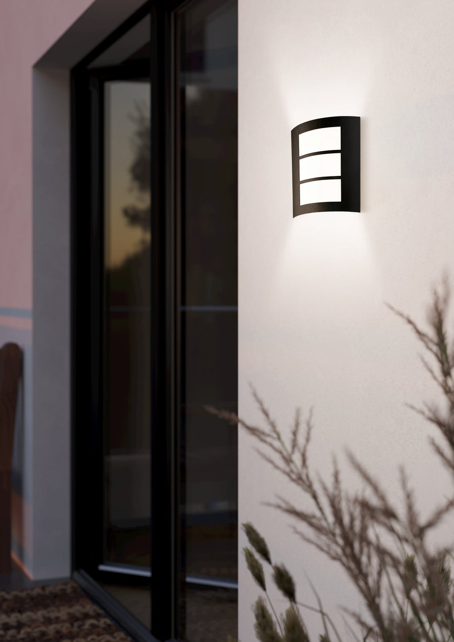 Applique extérieure noire allumée sur un mur blanc près d'une porte vitrée.