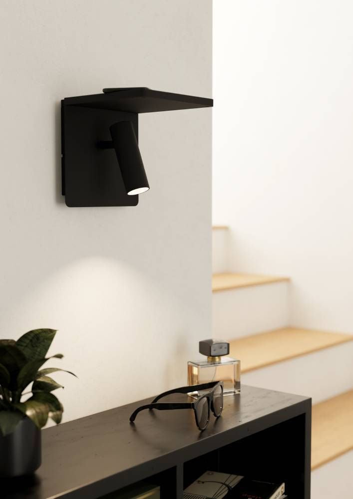 Applique noire allumée sur un mur blanc, au-dessus d'un meuble avec plante et lunettes.