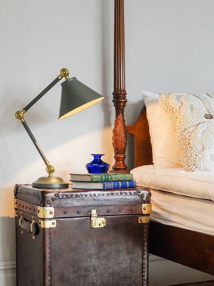 Lampe posée sur malle ancienne près du lit, avec livres et coussins clairs