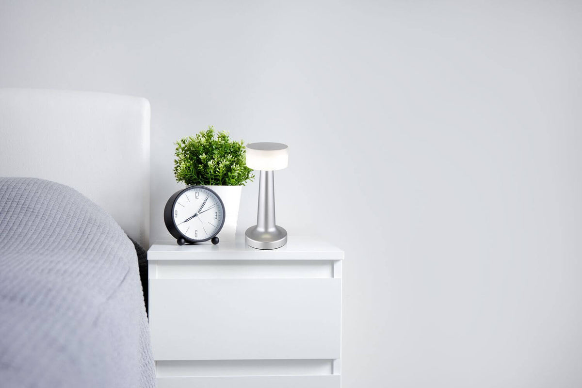 Mise en ambiance sur une table de chevet blanche avec réveil, plante et lit.
