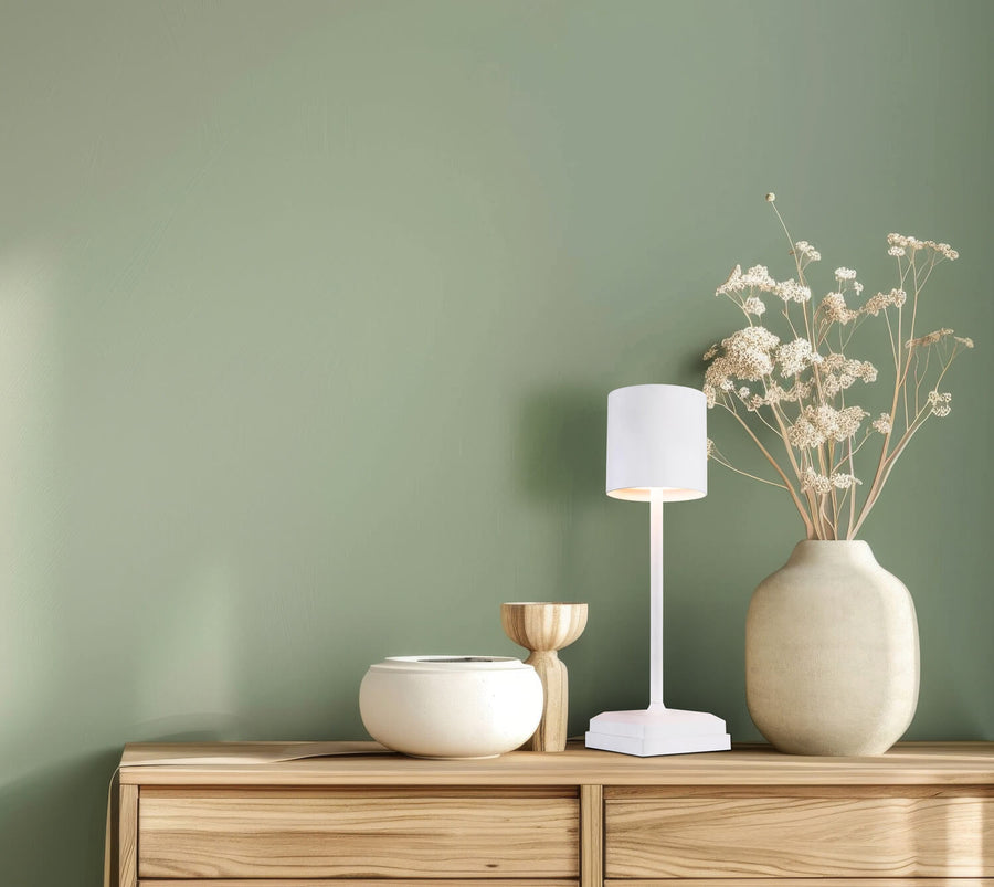 Lampe blanche allumée posée sur un meuble en bois devant un mur vert.