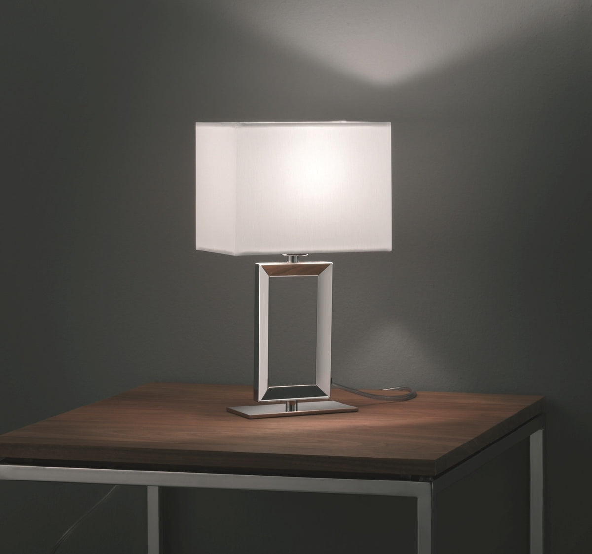 Lampe à poser allumée sur table en bois, abat-jour rectangulaire blanc et socle chromé.