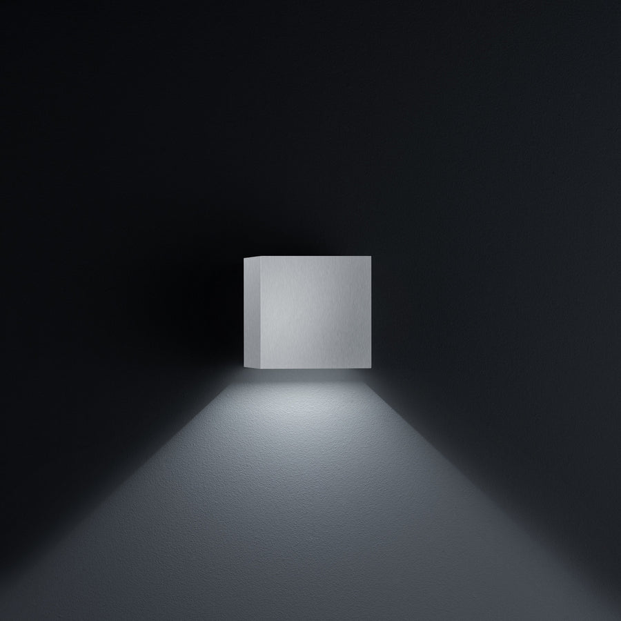 Applique cubique en aluminium brossé vue de face sur fond noir, halo vers le bas