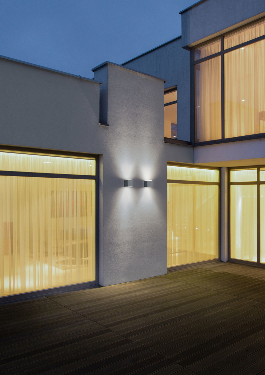 Mise en ambiance nocturne, deux appliques cubiques éclairent la façade d'une maison avec terrasse en bois.