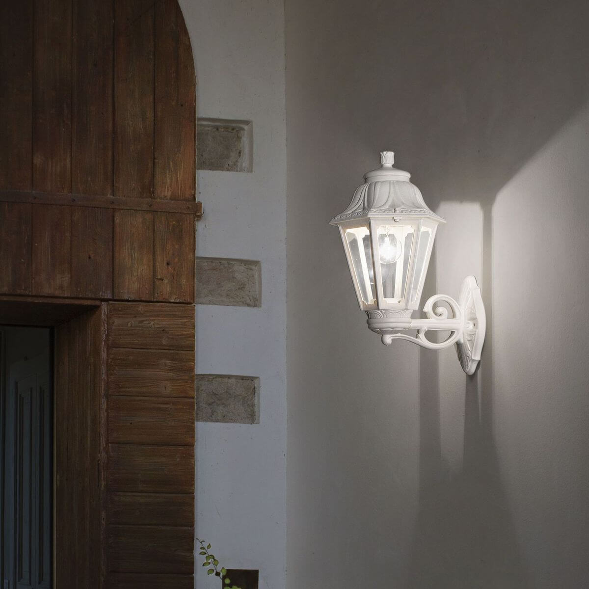 Applique lanterne blanche allumée, installée sur un mur extérieur devant une porte en bois