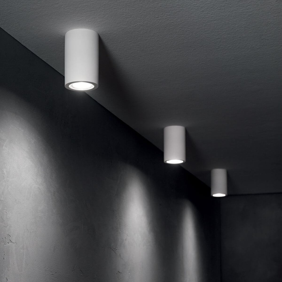 Trois plafonniers blancs au plafond sombre, ambiance béton minimaliste, faisceaux lumineux