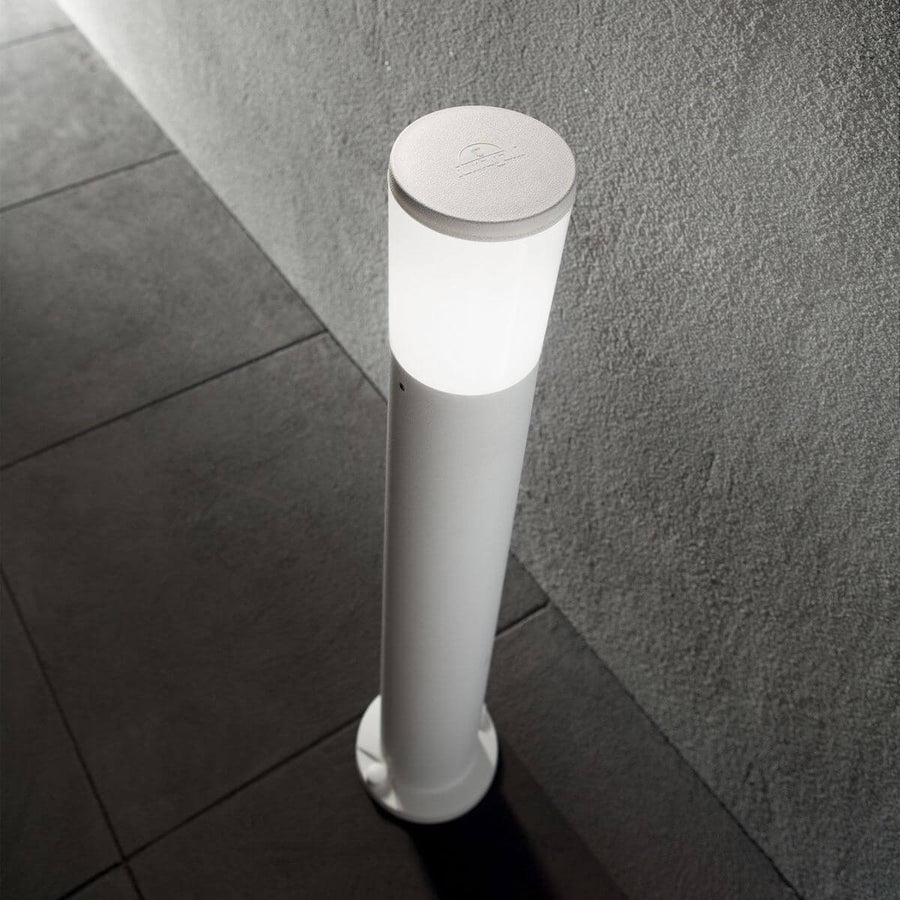 Lampadaire extérieur blanc allumé devant un mur gris, sur des dalles