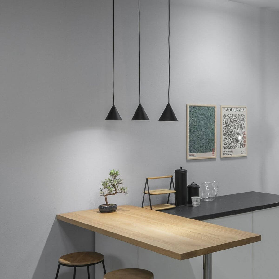 Trois suspensions noires coniques éclairant une table en bois dans un intérieur gris.