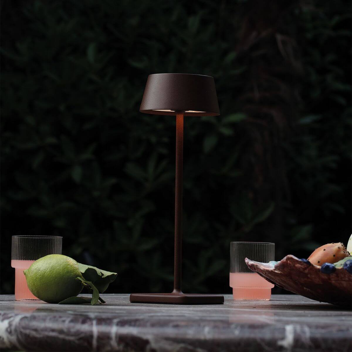 Mise en ambiance nocturne : lampe allumée sur table de marbre, verres, citron vert et feuillage sombre