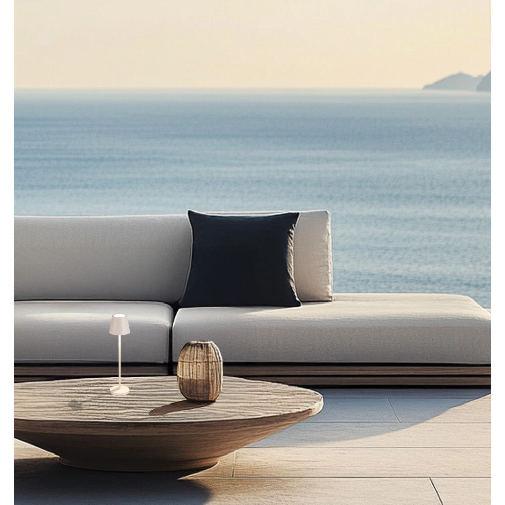 Petite lampe beige posée sur une table basse en extérieur devant un canapé et la mer.