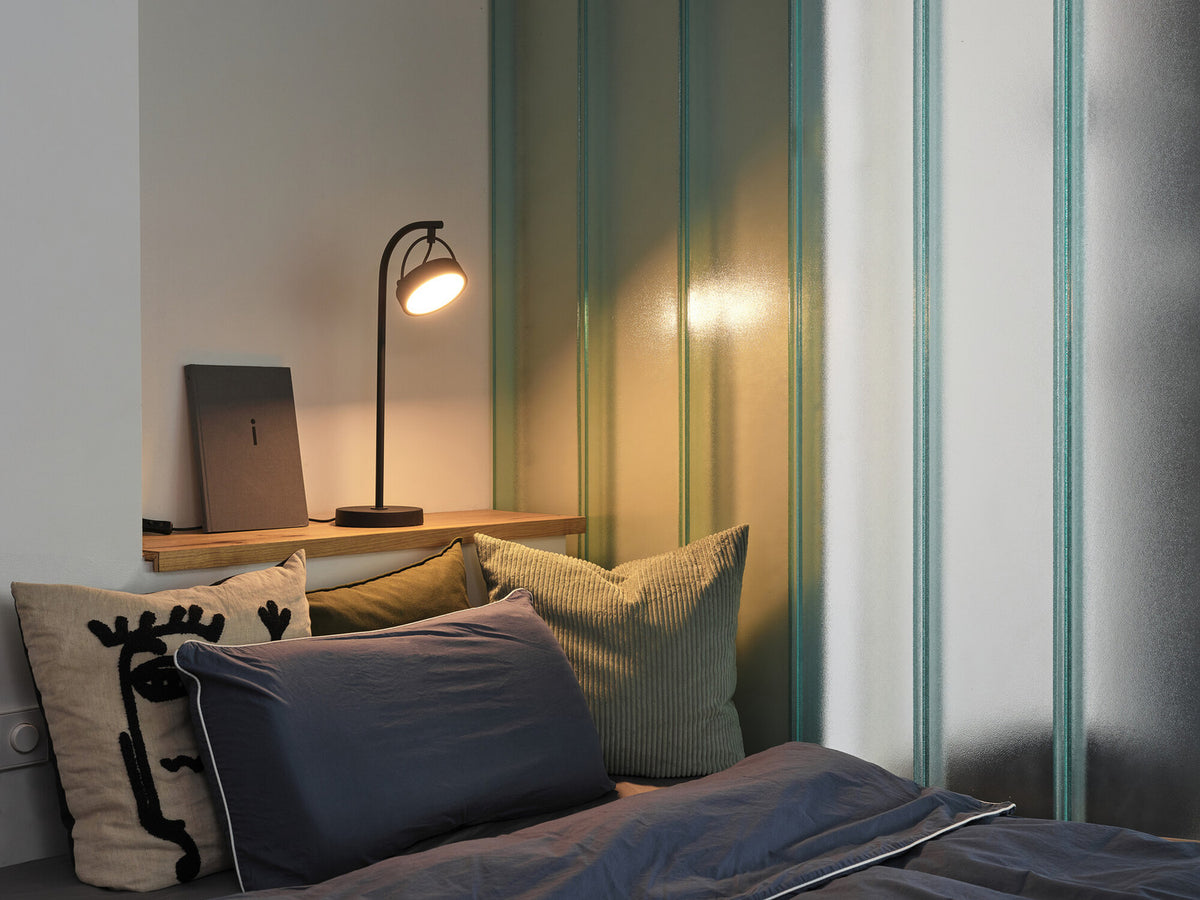 Lampe allumée sur une étagère de chevet, ambiance chambre cosy avec coussins.