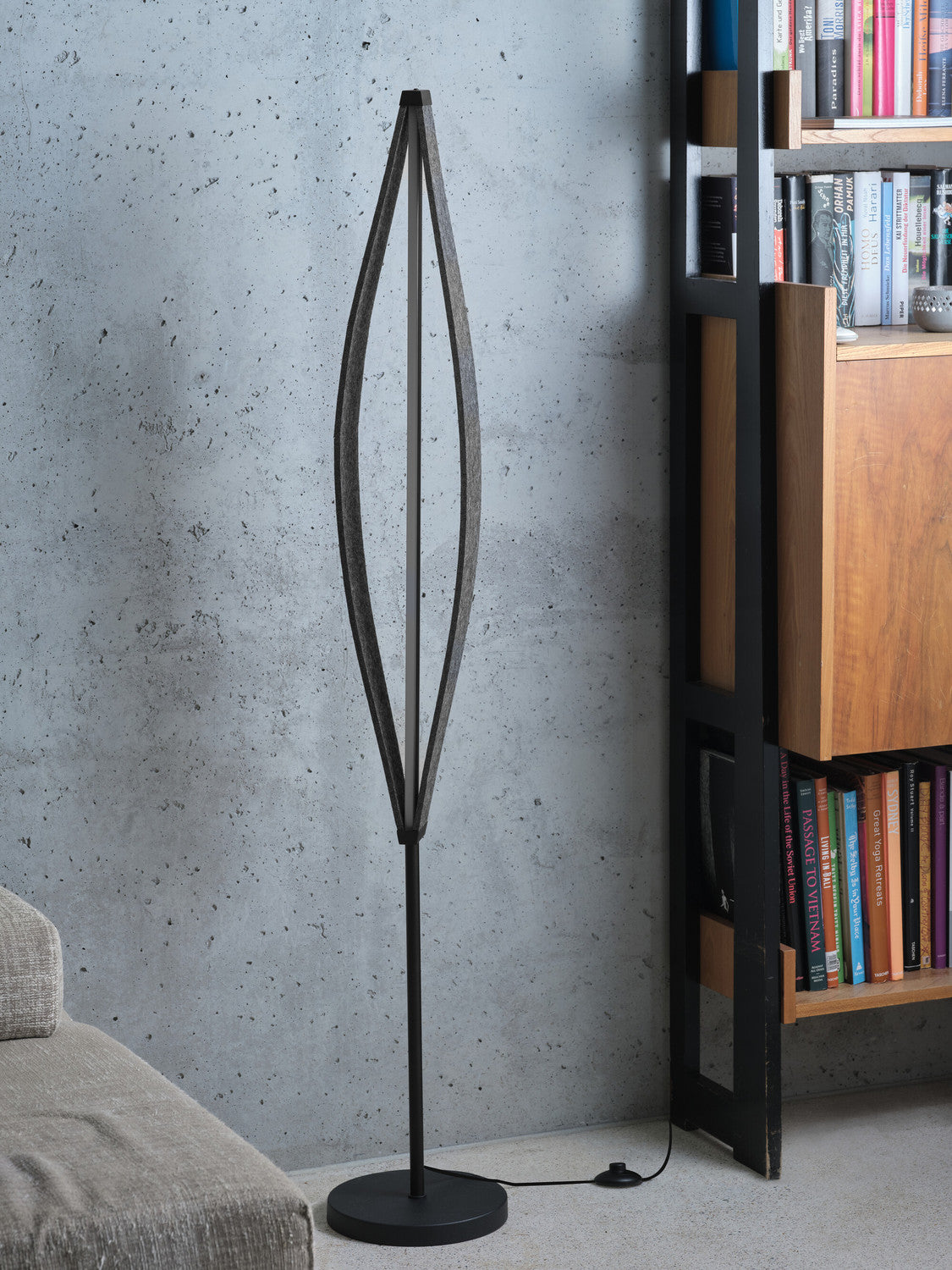Lampadaire anthracite éteint devant un mur béton, à côté d’une étagère en bois et d’un canapé.